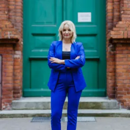 Profesjonalna kobieta w kobaltowym garniturze stoi przed zielonymi drzwiami z ceglaną fasadą, prezentując pewność siebie i kompetencje.