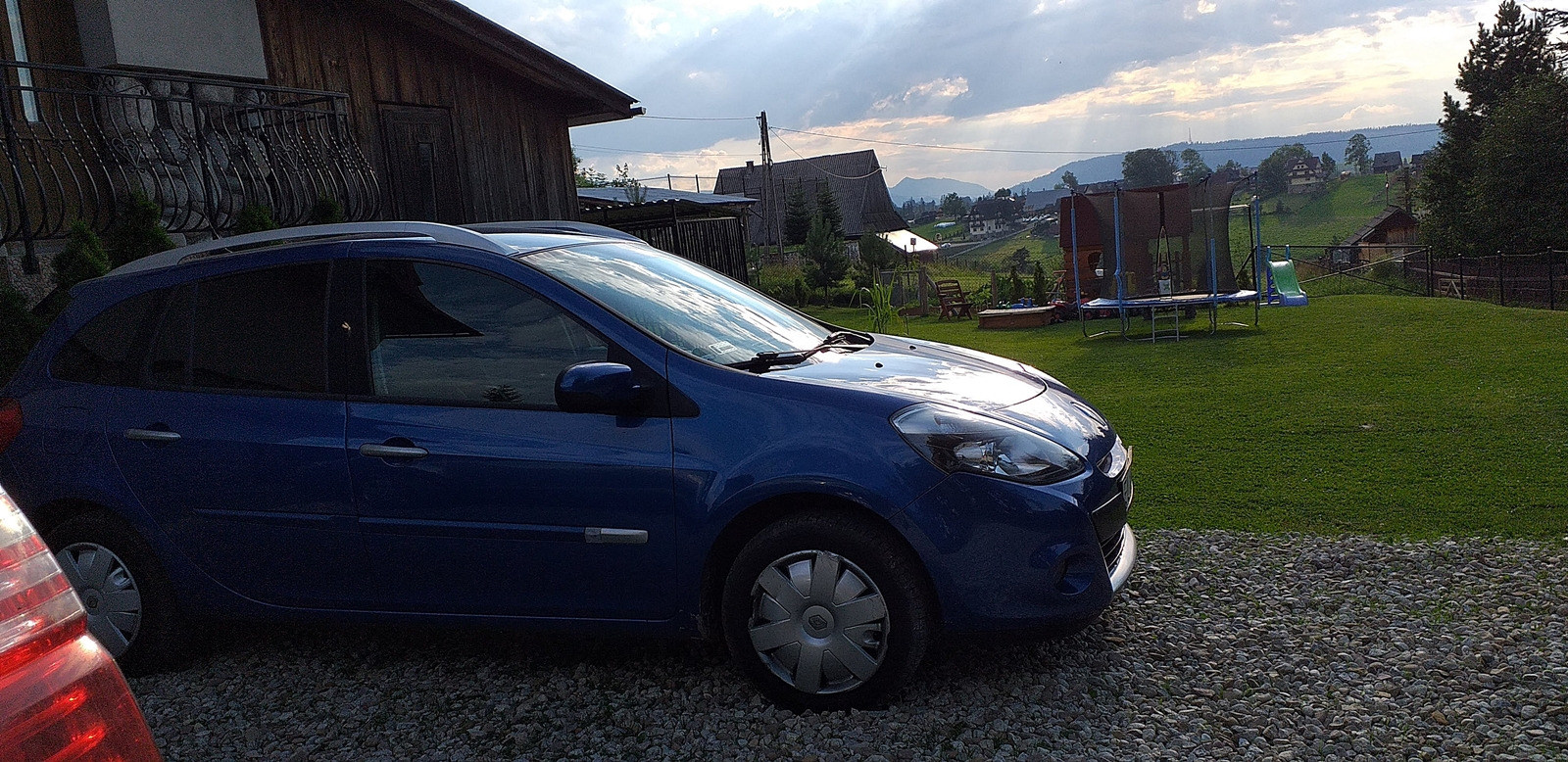 Niebieski samochód osobowy na tle zielonego ogrodu z trampoliną i widokiem na góry w oddali.