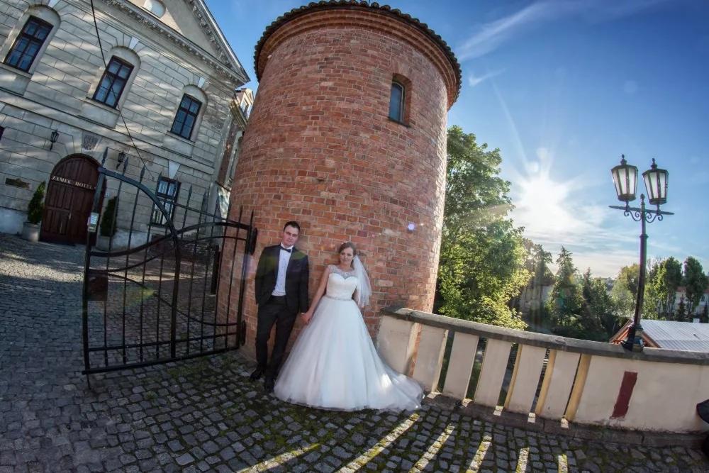 Para młoda pozuje przy ceglanej wieży i bramie w zabytkowym otoczeniu, słoneczny dzień.