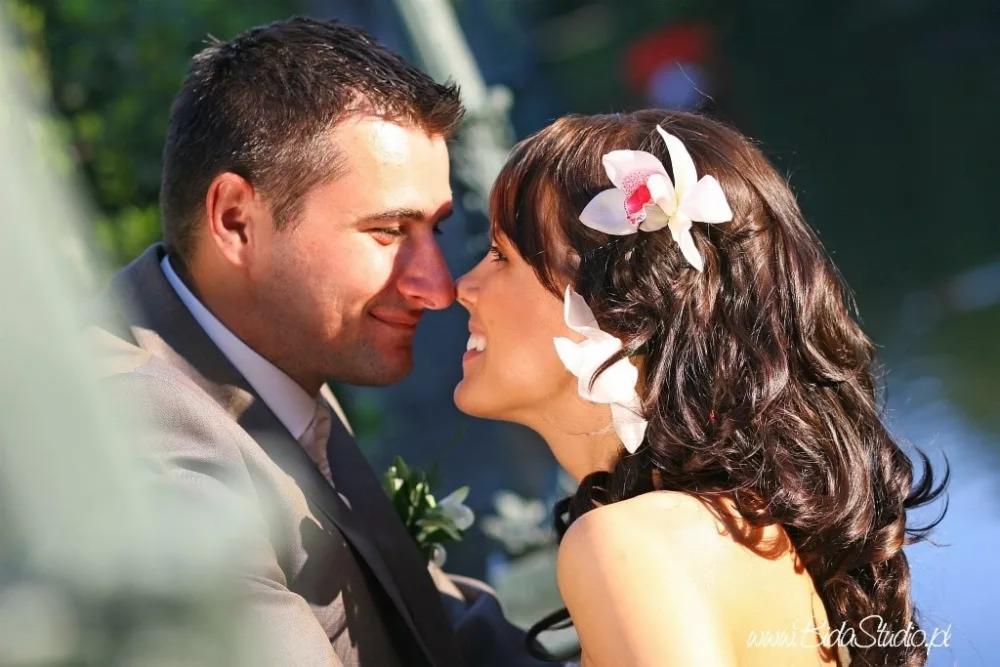 Uśmiechnięta para młoda, zbliżona twarzami, ona z białymi kwiatami we włosach, on w szarym garniturze, delikatne światło.