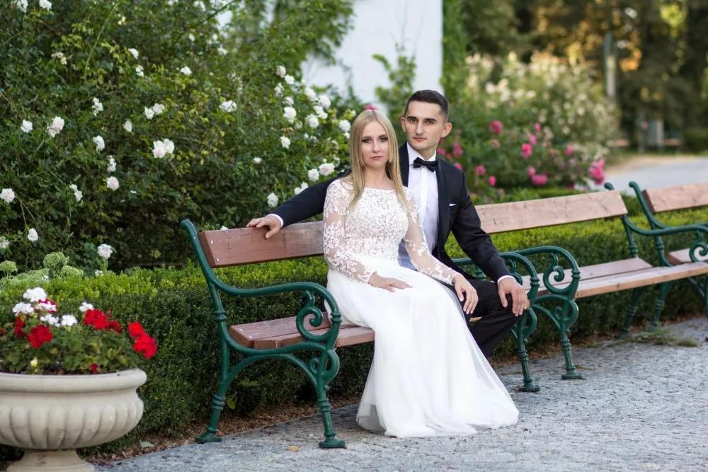 Para młoda siedzi na ławce w parku; panna młoda w koronkowej sukni z długim rękawem, pan młody w czarnym smokingu z muszką, w tle kwitnące krzewy róż.