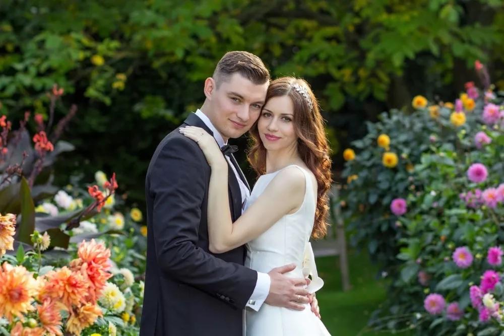 Para młoda w objęciach na tle kwitnącego ogrodu, panna młoda z ozdobą we włosach, pan młody w czarnym smokingu.