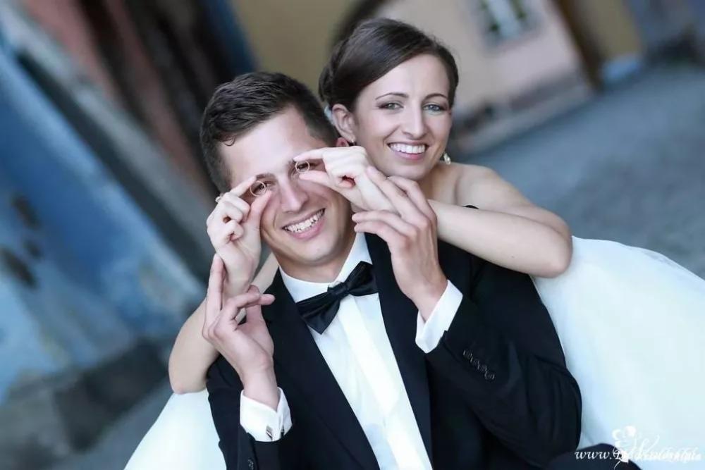 Uśmiechnięta para młoda w zbliżeniu, panna młoda obejmuje pana młodego, oboje patrzą przez obrączki trzymane przed oczami pana młodego.