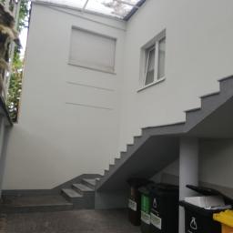 DOMASHOVETS - Budowa i remont od A do Z - Zewnętrzne schody z szarymi stopniami prowadzące na piętro, biała ściana z oknem i roletą, zadaszenie z przezroczystych płyt, obok pojemniki na śmieci z logo EKARO.