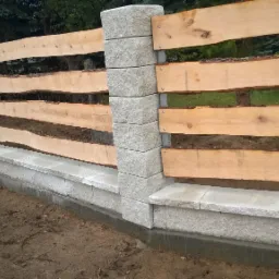 Nowoczesne ogrodzenie z surowego drewna i kamiennych słupków, z widoczną korą na deskach, osadzone na betonowej podmurówce.