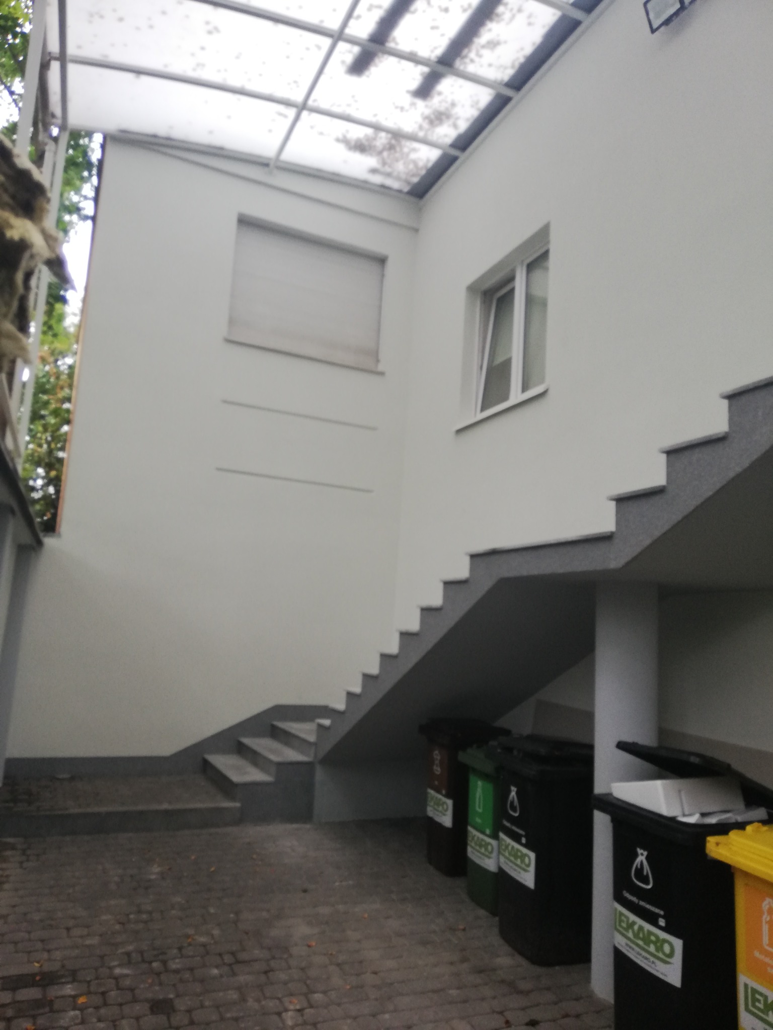 Zewnętrzne schody z szarymi stopniami prowadzące na piętro, biała ściana z oknem i roletą, zadaszenie z przezroczystych płyt, obok pojemniki na śmieci z logo EKARO.