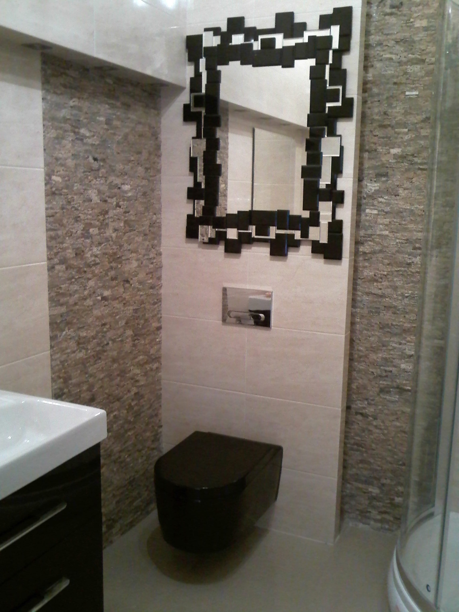 Nowoczesna łazienka z czarną toaletą i lustrem z geometryczną ramą, wykończona płytkami w odcieniach beżu i brązu, z widoczną kabiną prysznicową.