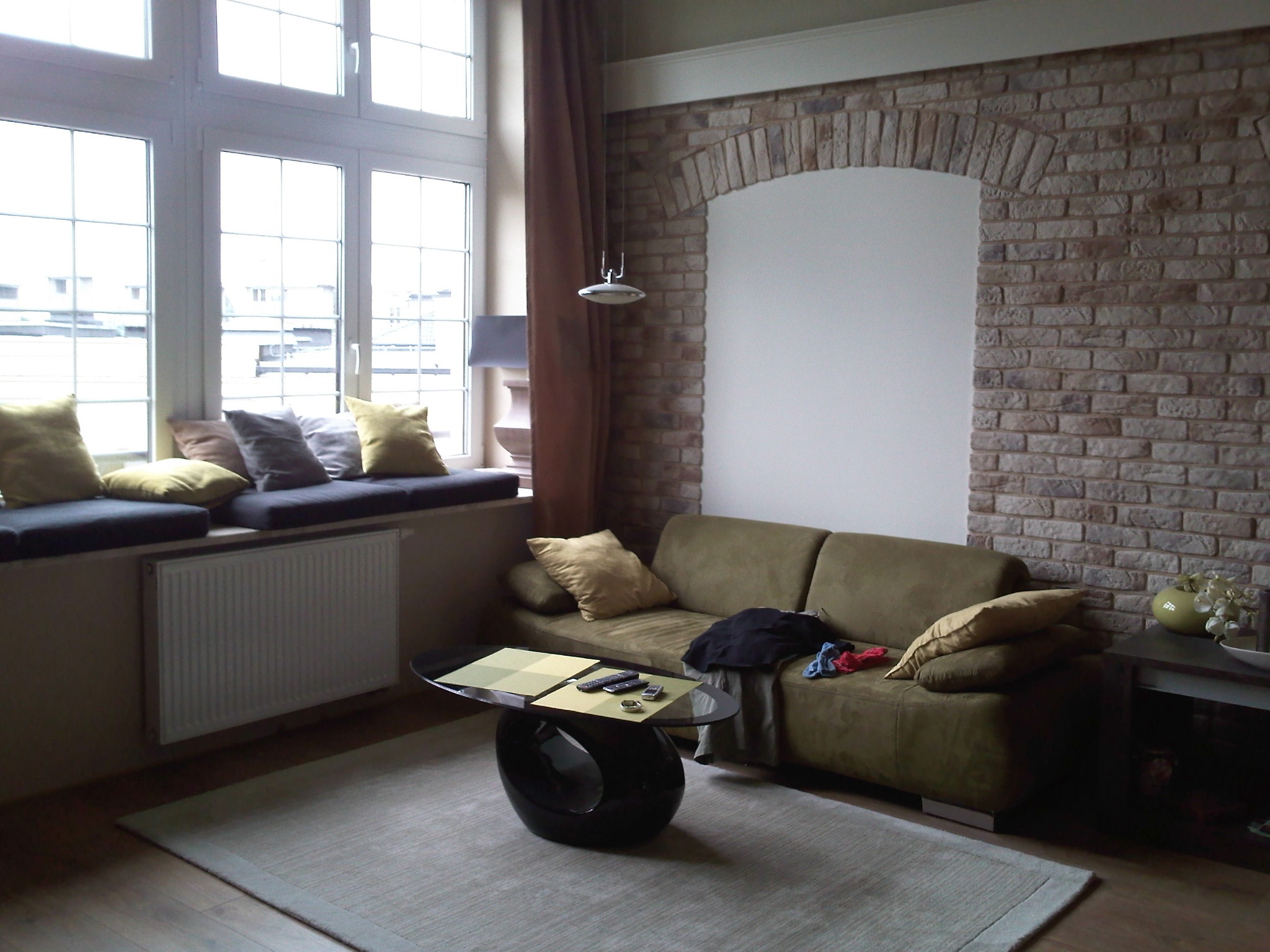 Jasne wnętrze pokoju dziennego z ceglaną ścianą z łukiem, dużym oknem z poduszkami, zieloną kanapą i nowoczesnym stolikiem kawowym.