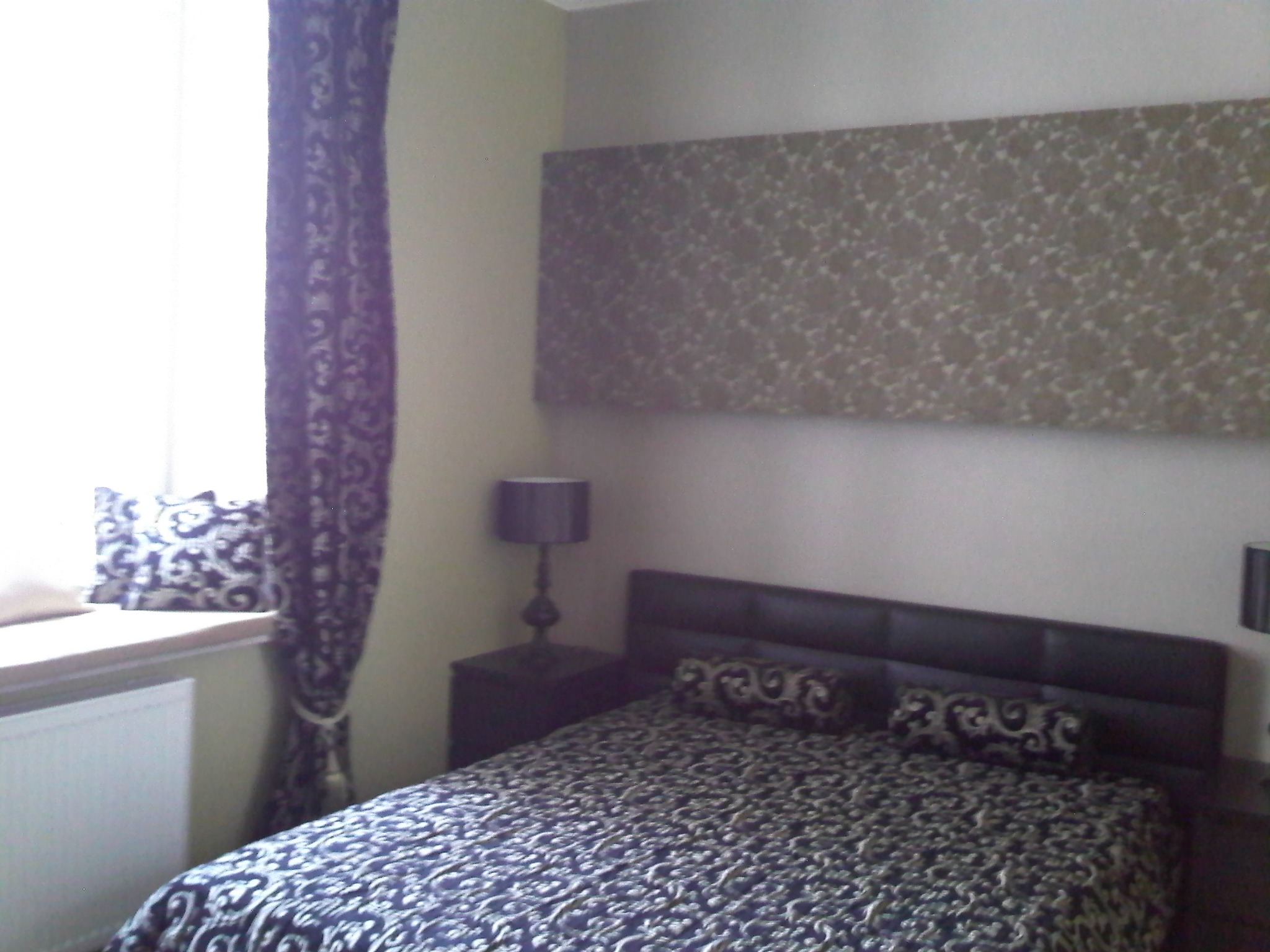 Sypialnia z wzorzystą pościelą i zasłonami w odcieniach szarości i fioletu, lampka nocna na szafce, dekoracyjny panel nad łóżkiem.