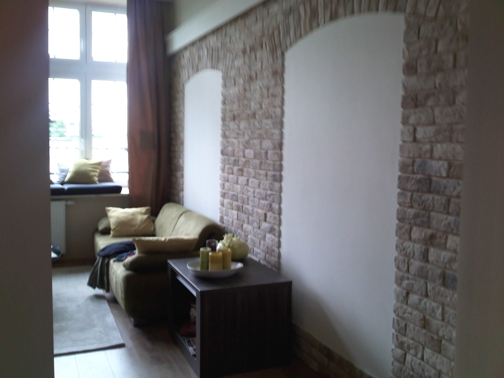 Salon z dekoracyjną ścianą z cegły imitującej kamień, wnękami w kształcie łuków, zieloną sofą i stolikiem z dekoracjami.