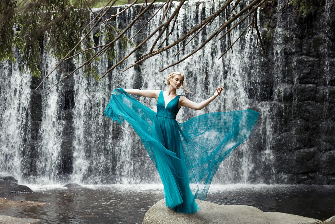 Kobieta w turkusowej sukni na tle wodospadu, pod zwisającymi gałęziami, pozuje z rozłożonymi ramionami.