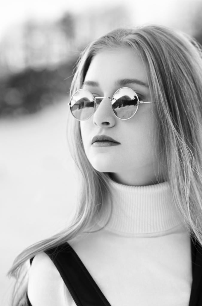 Portret młodej kobiety w okrągłych okularach, odbijających krajobraz, w czarno-białej tonacji, z delikatnym uśmiechem i długimi włosami.