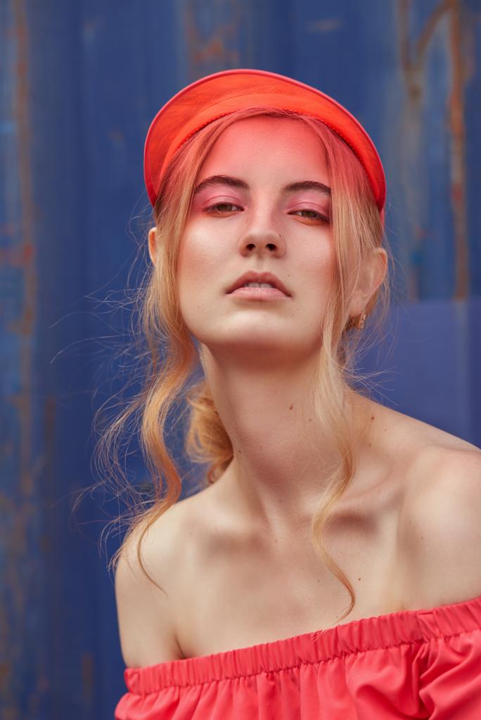 Portret kobiety w czerwonym kapeluszu i bluzce na tle niebieskiej ściany. Subtelny makijaż i delikatne światło podkreślają jej urodę. Ujęcie od dołu.