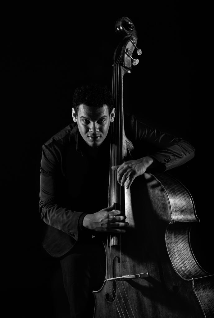 Czarno-białe zdjęcie muzyka z kontrabasem w ciemnym otoczeniu, artystyczne oświetlenie podkreśla teksturę instrumentu i wyraz twarzy.