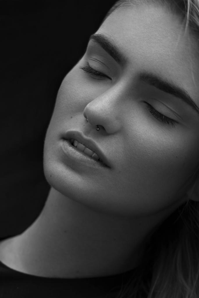 Portret kobiety z kolczykiem w nosie, zamknięte oczy, czarno-białe zdjęcie, delikatny makijaż, spokojny wyraz twarzy, ciemne tło.