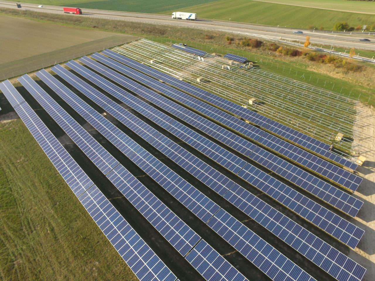 Widok z góry na dużą farmę paneli słonecznych, część paneli już zamontowana, a część konstrukcji montażowej jeszcze czeka na instalację, w tle autostrada z jadącymi samochodami.