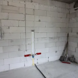 Wewnętrzna instalacja elektryczna w surowym pomieszczeniu z betonową podłogą i ścianami z białych bloczków, widoczne kable w listwach, puszki elektryczne oraz fragment instalacji wodnej...