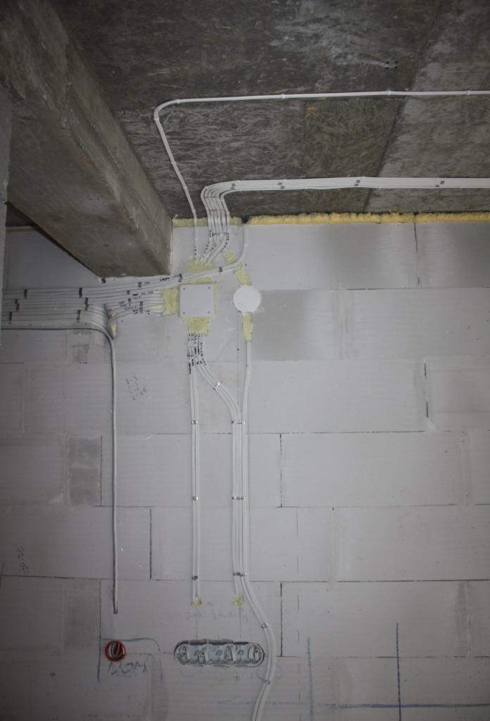 Białe przewody elektryczne prowadzone po ścianie z bloczków betonowych, zabezpieczone obejmami, z widoczną puszką rozgałęźną i fragmentem sufitu z betonową belką.