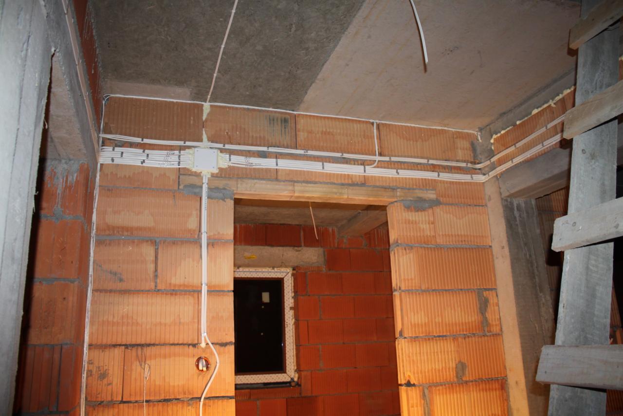 Wewnętrzna instalacja elektryczna w budynku w trakcie budowy: białe przewody ułożone wzdłuż ceglanej ściany, puszka elektryczna, widoczny fragment okna i drewniane rusztowanie.