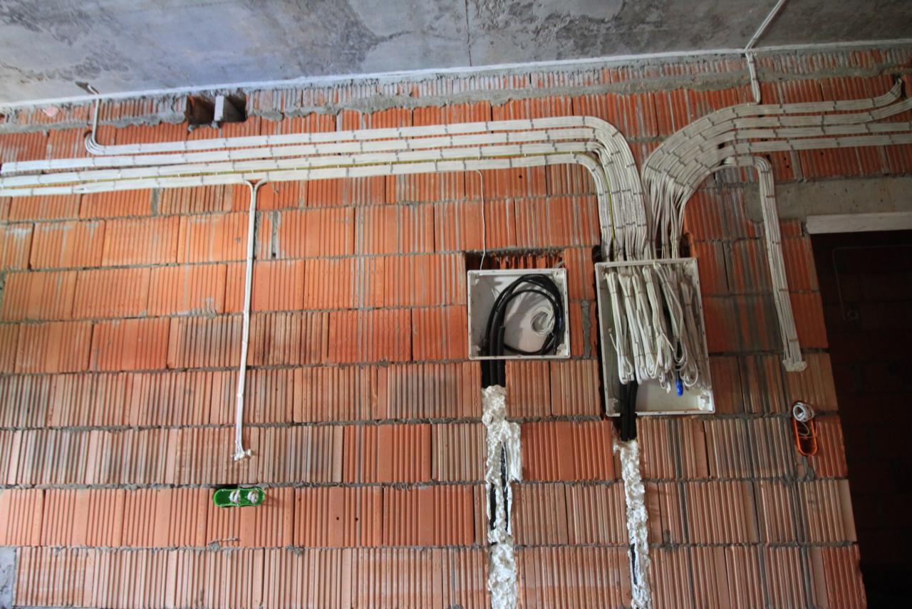 Instalacja elektryczna w trakcie montażu na ścianie z cegły, widoczne kable w peszlach, puszki instalacyjne z przewodami oraz pianka montażowa w bruzdach.