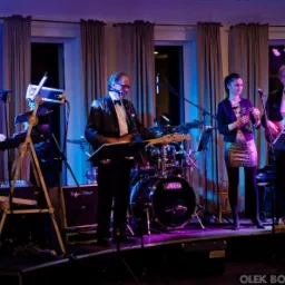 Zespół muzyczny na scenie w trakcie występu, mężczyźni w błyszczących marynarkach, kobieta z tamburynem, perkusja Yamaha w tle.