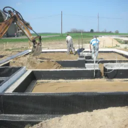 Budowa fundamentów domu: wykop z izolacją przeciwwilgociową, pracownicy i koparka w tle. Widoczne szalunki i przygotowanie do wylewki.