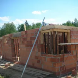 Wznoszenie ścian domu z czerwonej cegły ceramicznej. Widoczne drewniane szalunki okienne i zbrojenie. Niebieskie niebo w tle. Prace budowlane w toku.