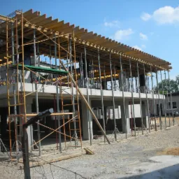 Dwukondygnacyjny budynek w trakcie budowy, betonowy szkielet z drewnianym szalunkiem stropu, podpory pionowe, rusztowania, żwirowe podłoże, błękitne niebo.