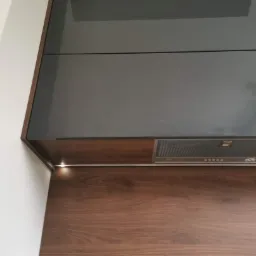Kuchenny okap podszafkowy z oświetleniem LED, zamontowany pod ciemnoszarymi szafkami, na tle drewnianej ściany.