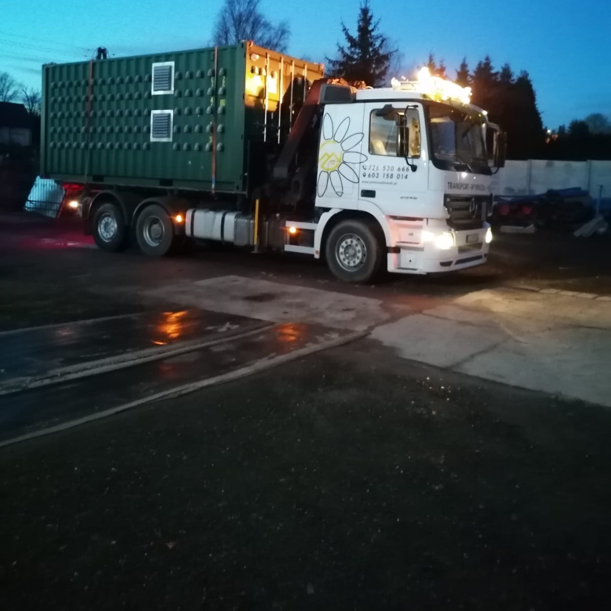 Ciężarówka z naczepą kontenerową w kolorze khaki, z zamontowanym żurawiem, zaparkowana na placu budowy o zmierzchu, oświetlona reflektorami, z widocznym logo firmy transportowej na kabinie.