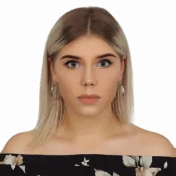 Portret młodej kobiety z blond włosami i kolczykami kołami, ubrana w bluzkę z odkrytymi ramionami w kwiatowy wzór, na białym tle.