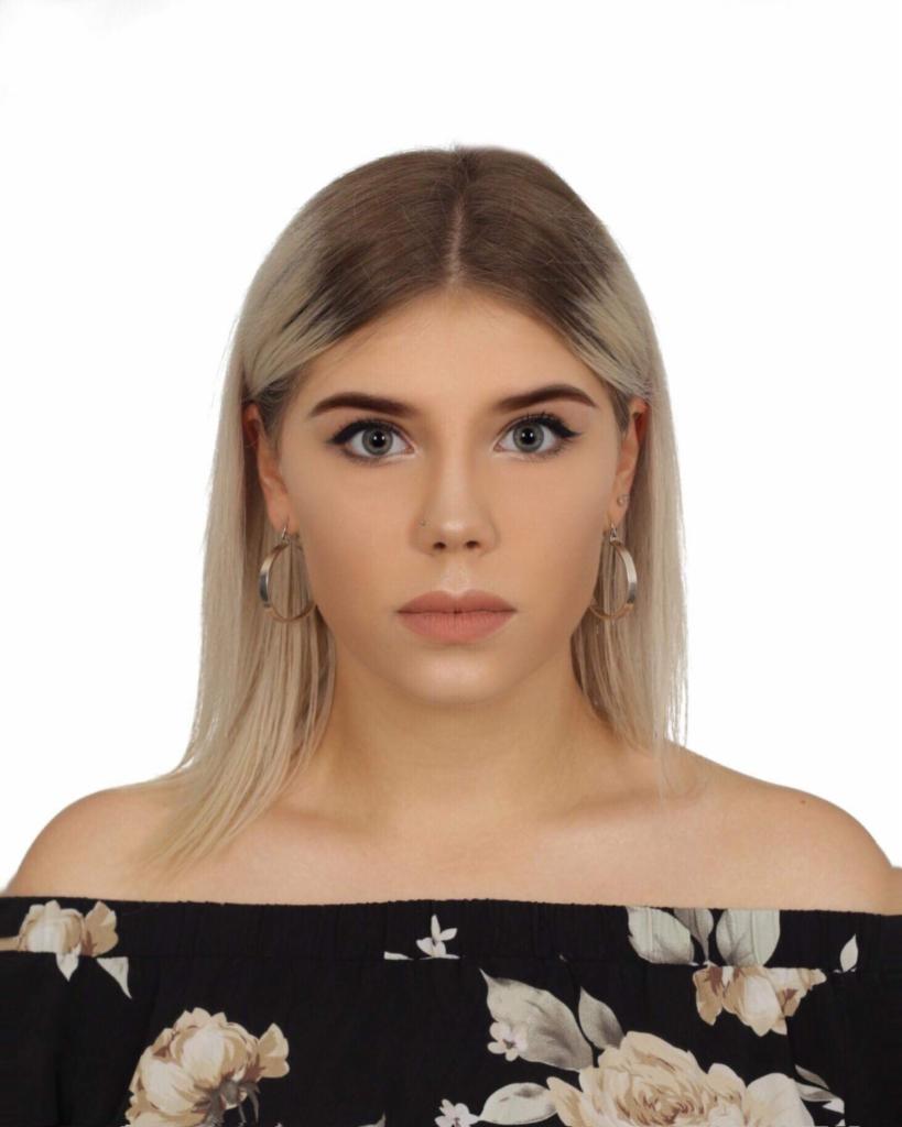 Portret młodej kobiety z blond włosami i kolczykami kołami, ubrana w bluzkę z odkrytymi ramionami w kwiatowy wzór, na białym tle.