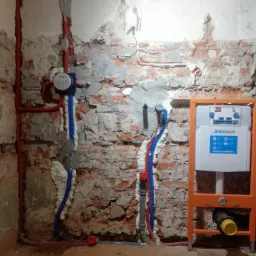 Surowa ściana z cegły i betonu, w trakcie montażu instalacji wodno-kanalizacyjnej z rurami w izolacji, widoczny stelaż podtynkowy WC JOMOTech i licznik wody Grohe.