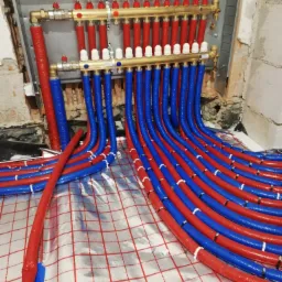 Rozdzielacz ogrzewania podłogowego z rurami w kolorze czerwonym i niebieskim, ułożonymi na folii z nadrukowaną siatką, widoczne fragmenty ściany w stanie surowym.