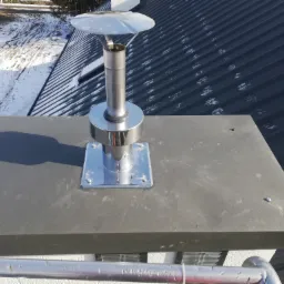 Komin wentylacyjny ze stali nierdzewnej na dachu pokrytym blachą trapezową; widoczne ślady śniegu na dachu i w tle.