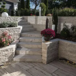 Schody zewnętrzne i murki oporowe z jasnego kamienia, otoczone roślinnością i ozdobnymi kulami, z widocznymi przewodami na ziemi.