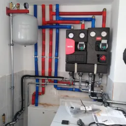 Kompletna instalacja centralnego ogrzewania z piecem, zbiornikiem wyrównawczym, pompami obiegowymi i rurami w kolorach czerwonym i niebieskim, zamontowana na ścianie wyłożonej jasnymi płytkami...