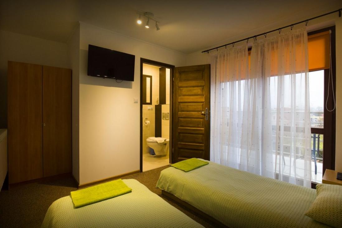 Pokój hotelowy z dwoma łóżkami pojedynczymi, widok na otwarte drzwi do łazienki z toaletą i umywalką, oraz okno balkonowe z białymi zasłonami i pomarańczową roletą.