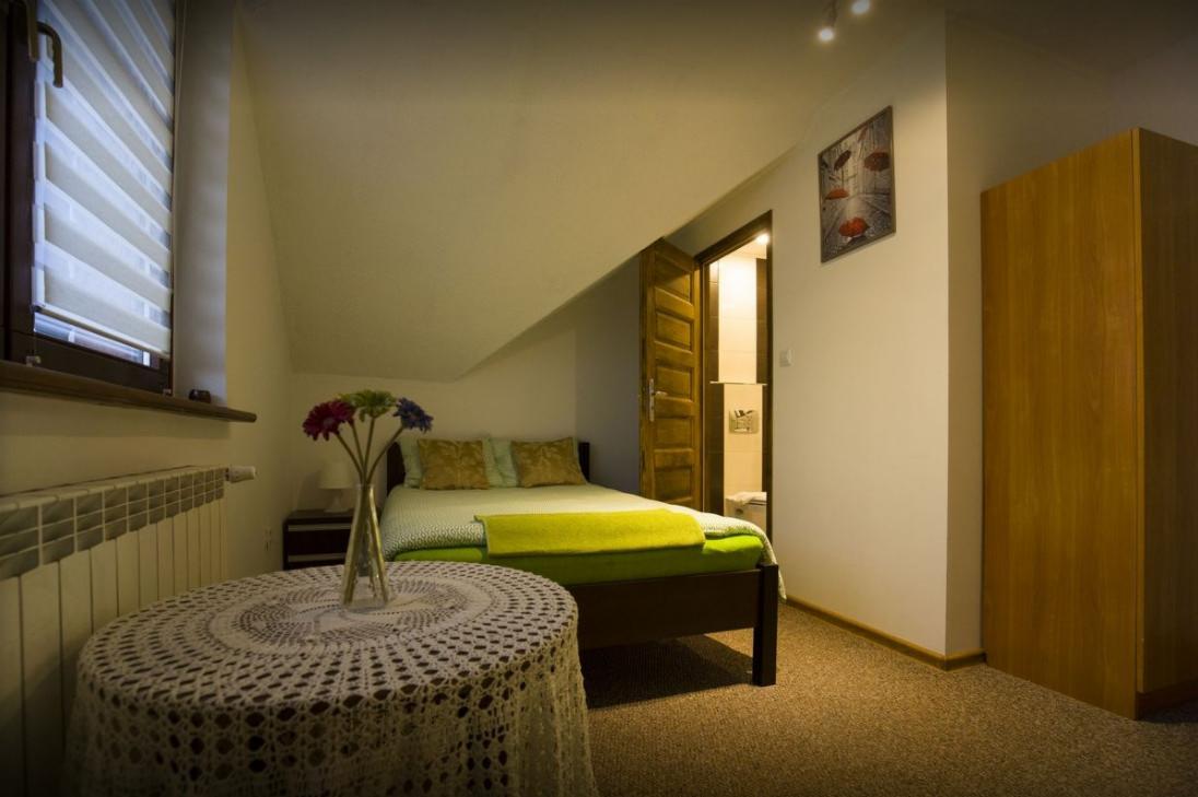 Pokój z łóżkiem, stolikiem z koronkowym obrusem i wazonem z kwiatami, z otwartymi drzwiami do łazienki w tle.