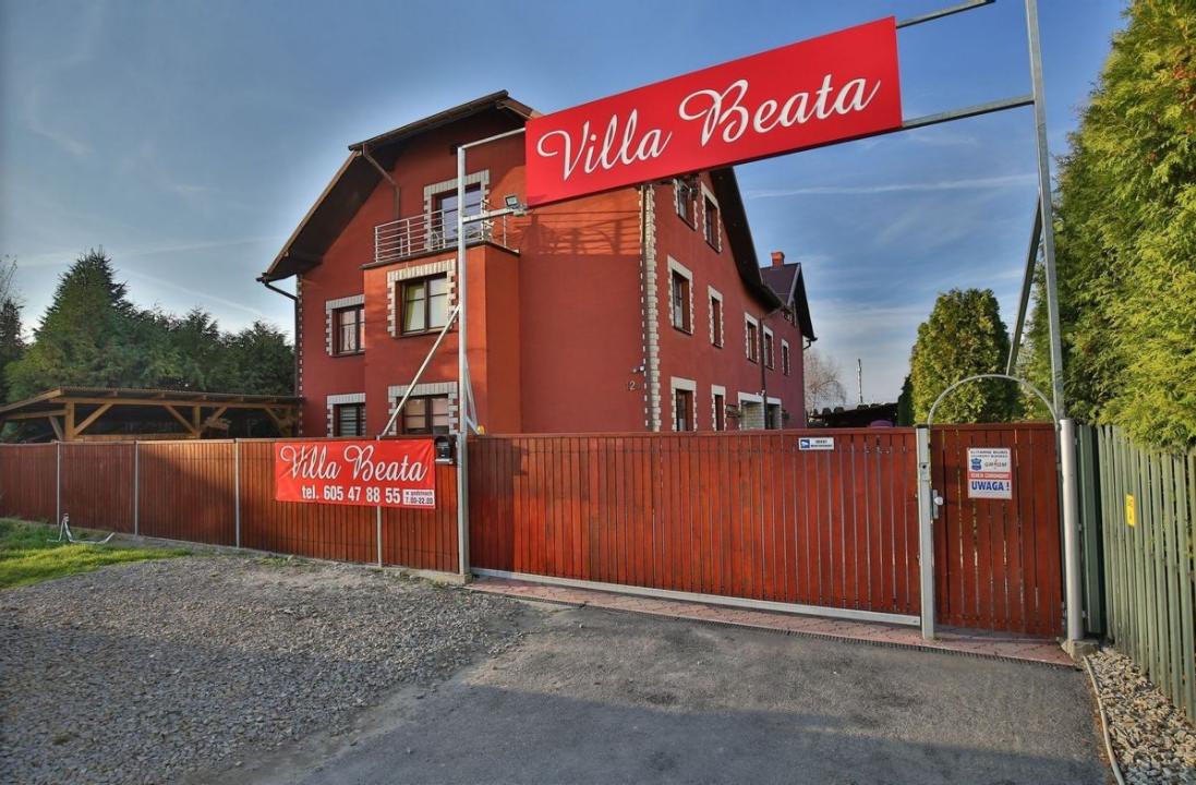 Czerwony budynek z brązowym ogrodzeniem i szyldem 'Villa Beata' nad wejściem, widok z zewnątrz w słoneczny dzień.