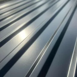 Szare, metalowe panele dachowe z wyraźnymi przetłoczeniami, ułożone ukośnie, w bliskim kadrze, z widocznym odbiciem światła.