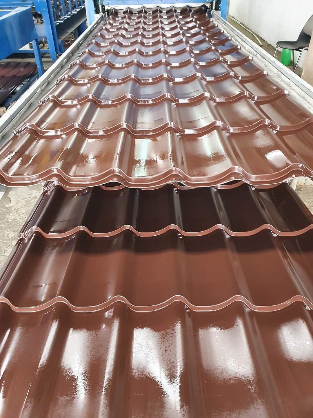Nowe, błyszczące arkusze brązowej blachy dachowej o falistym profilu, ułożone jeden na drugim w fabryce, w tle widoczna linia produkcyjna.