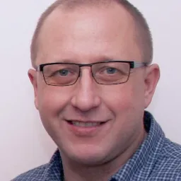 Portret mężczyzny w okularach i koszuli w kratę, uśmiechającego się do kamery na jasnym tle. Widoczne zmarszczki mimiczne i krótko ostrzyżone włosy.