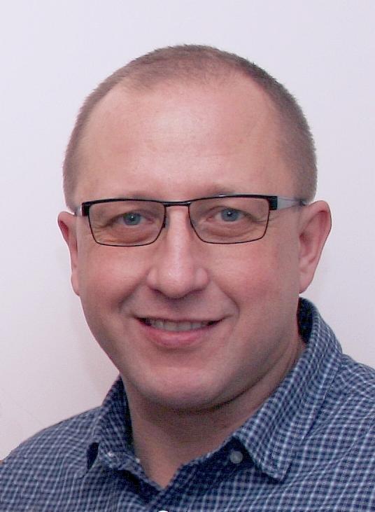 Portret mężczyzny w okularach i koszuli w kratę, uśmiechającego się do kamery na jasnym tle. Widoczne zmarszczki mimiczne i krótko ostrzyżone włosy.