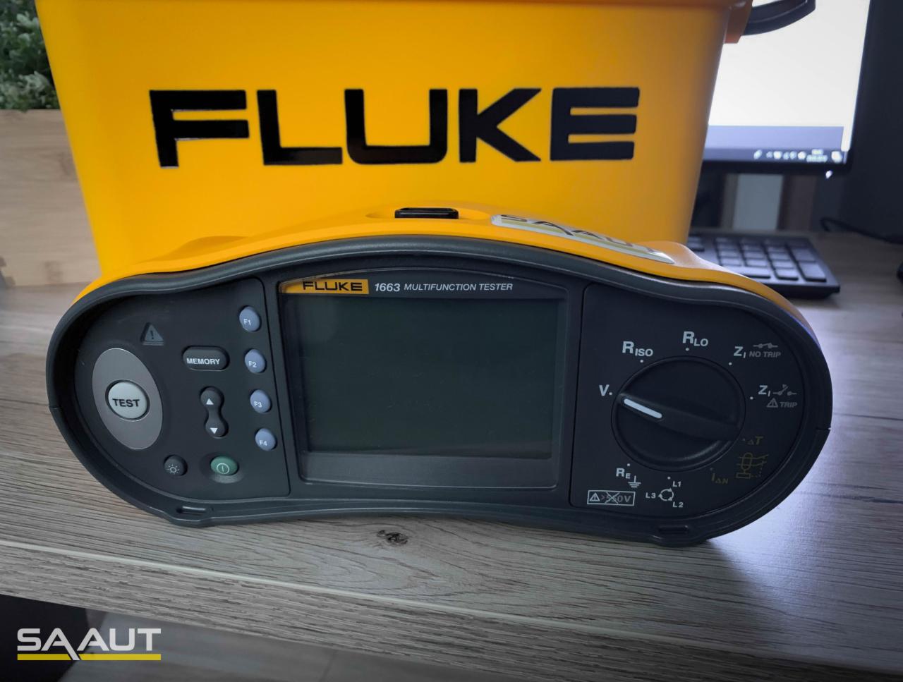 Multifunkcyjny tester instalacji elektrycznych Fluke 1663 z żółtym pojemnikiem na tle biurka z komputerem.