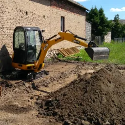 Żółta mini koparka JCB przygotowuje wykop pod fundamenty budynku z cegły, widoczne wytyczone linie i pryzma ziemi.