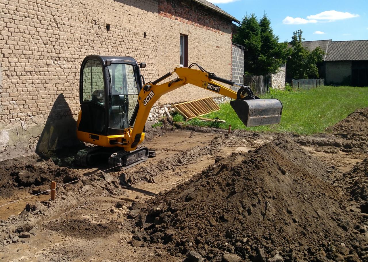 Żółta mini koparka JCB przygotowuje wykop pod fundamenty budynku z cegły, widoczne wytyczone linie i pryzma ziemi.