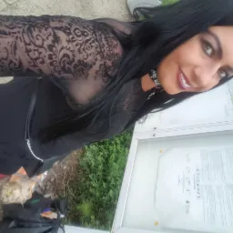 Uśmiechnięta kobieta w czarnej koronkowej bluzce pozuje obok tablicy informacyjnej. Widoczna zieleń w tle i fragment chodnika. Kadr z lekkiego ukosa.