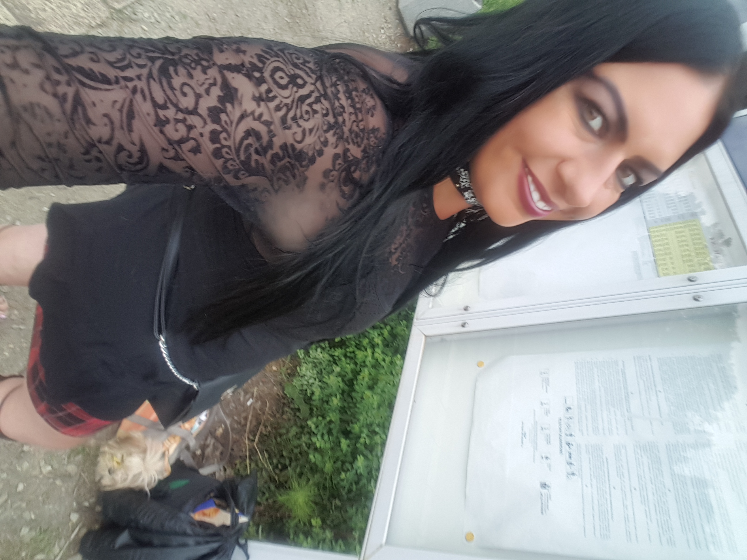 Uśmiechnięta kobieta w czarnej koronkowej bluzce pozuje obok tablicy informacyjnej. Widoczna zieleń w tle i fragment chodnika. Kadr z lekkiego ukosa.