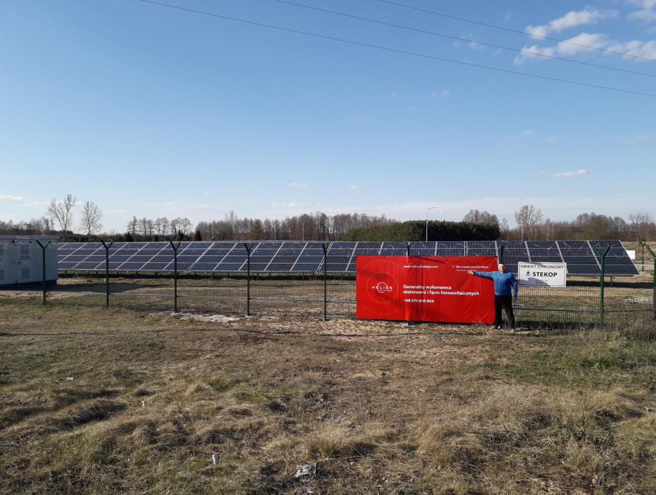 Instalacja fotowoltaiczna na farmie, widoczny rząd paneli za ogrodzeniem z siatki, na pierwszym planie mężczyzna wskazujący na baner firmy Helios, wykonawcy elektrowni i farm fotowoltaicznych.